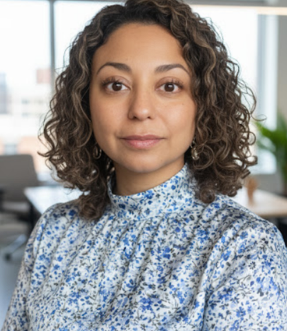 A woman with curly hair wearing a floral dress shirt, standing in an office environment with large windows in the background.