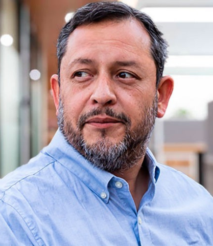 A middle-aged man with a beard, wearing a light blue shirt, looking thoughtfully to the side in an outdoor setting.